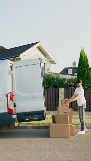 A man wearing casual clothing, including a white t-shirt and jeans, is standing outside next to a white moving van with its rear doors open. He is in the process of lifting or placing a cardboard cart