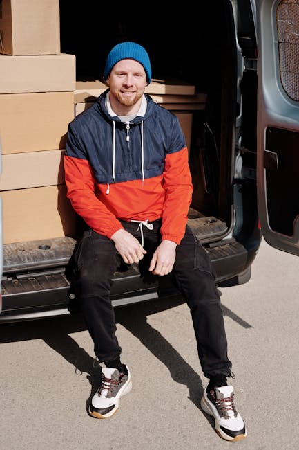 A man wearing casual clothing, including a white t-shirt and jeans, is standing outside next to a white moving van with its rear doors open. He is in the process of lifting or placing a cardboard cart