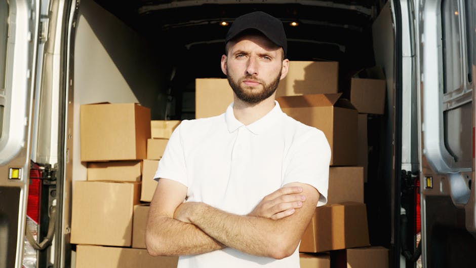 A man wearing casual clothing and glasses is standing beside an open white moving van parked on a residential street. He is holding a cardboard box, preparing to load it onto the vehicle. Several othe
