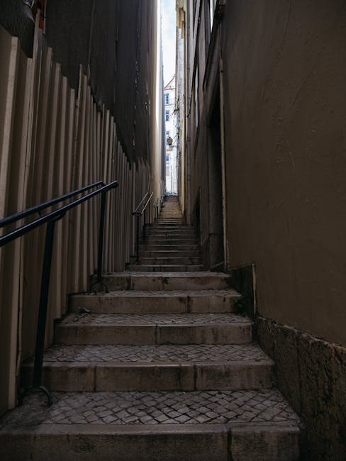 A narrow flight of outdoor concrete stairs with dark grey treads ascending between two beige building walls, which have visible piping, electrical meters, and small patches of vegetation. The surround