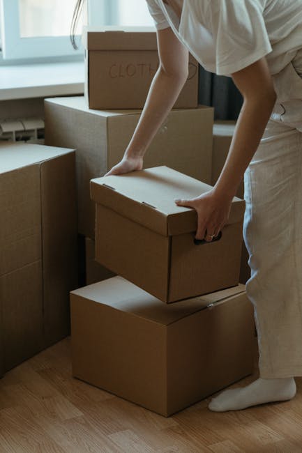 A man wearing a striped t-shirt and blue work overalls is sitting at the open side door of a white delivery van during a home relocation, handling paperwork with a pen. He is surrounded by cardboard b